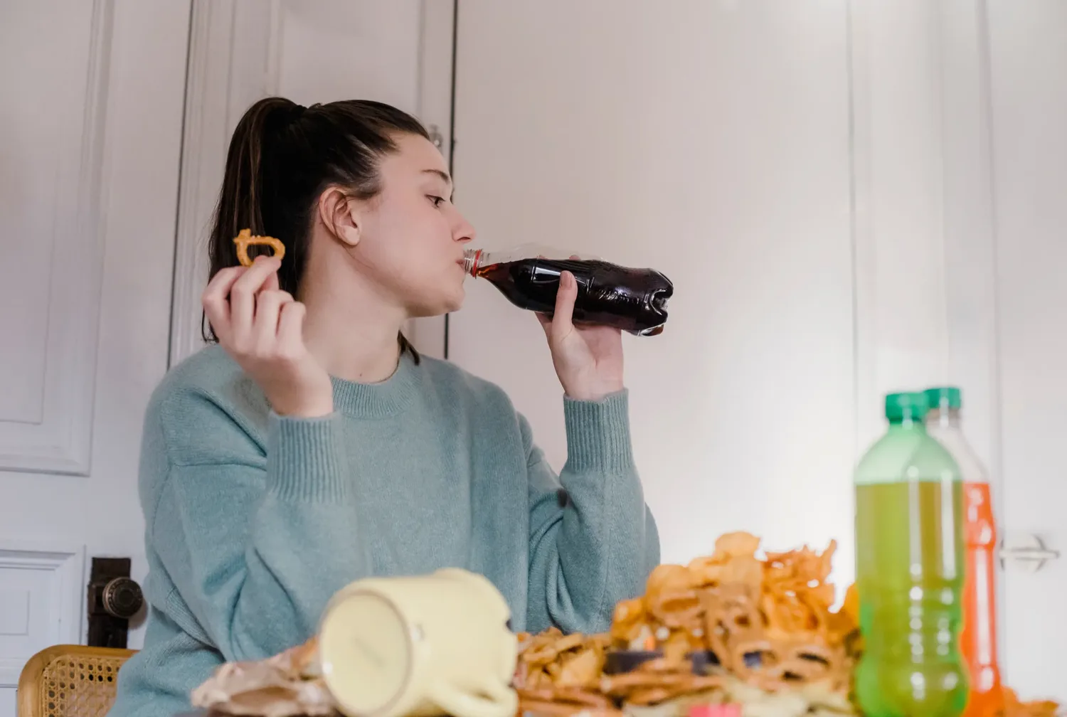 A young woman drinking a soft drink at a table covered with open snack packets and bottles.