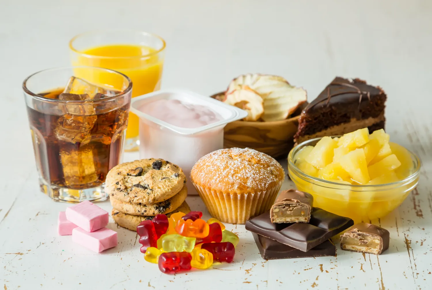 An assortment of sweet foods and drinks arranged on a light wooden surface, including cake, biscuits, chocolate, fruit and soft drinks.